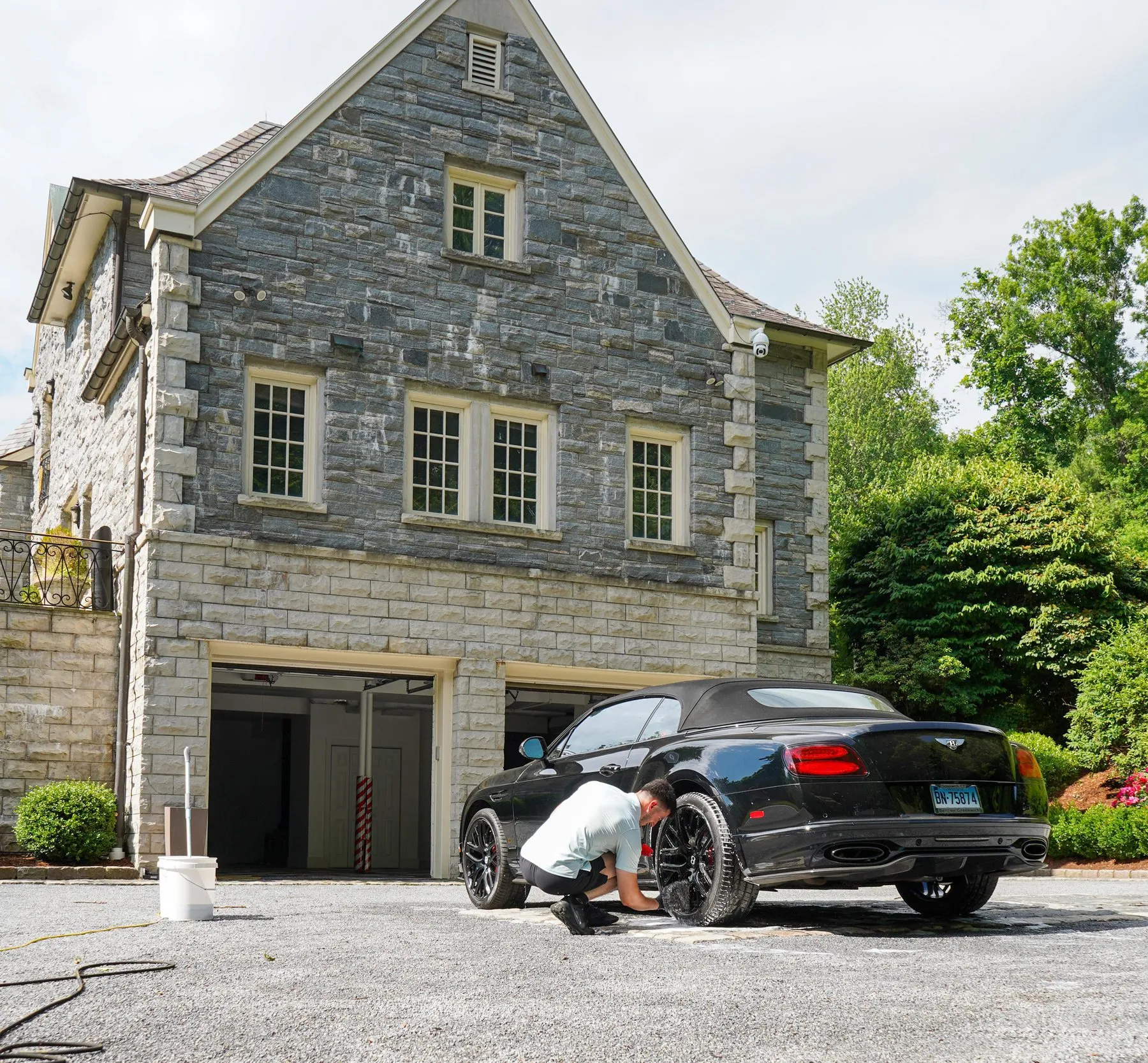 A detailer working on a black Bentley convertible in front of a large stone house.