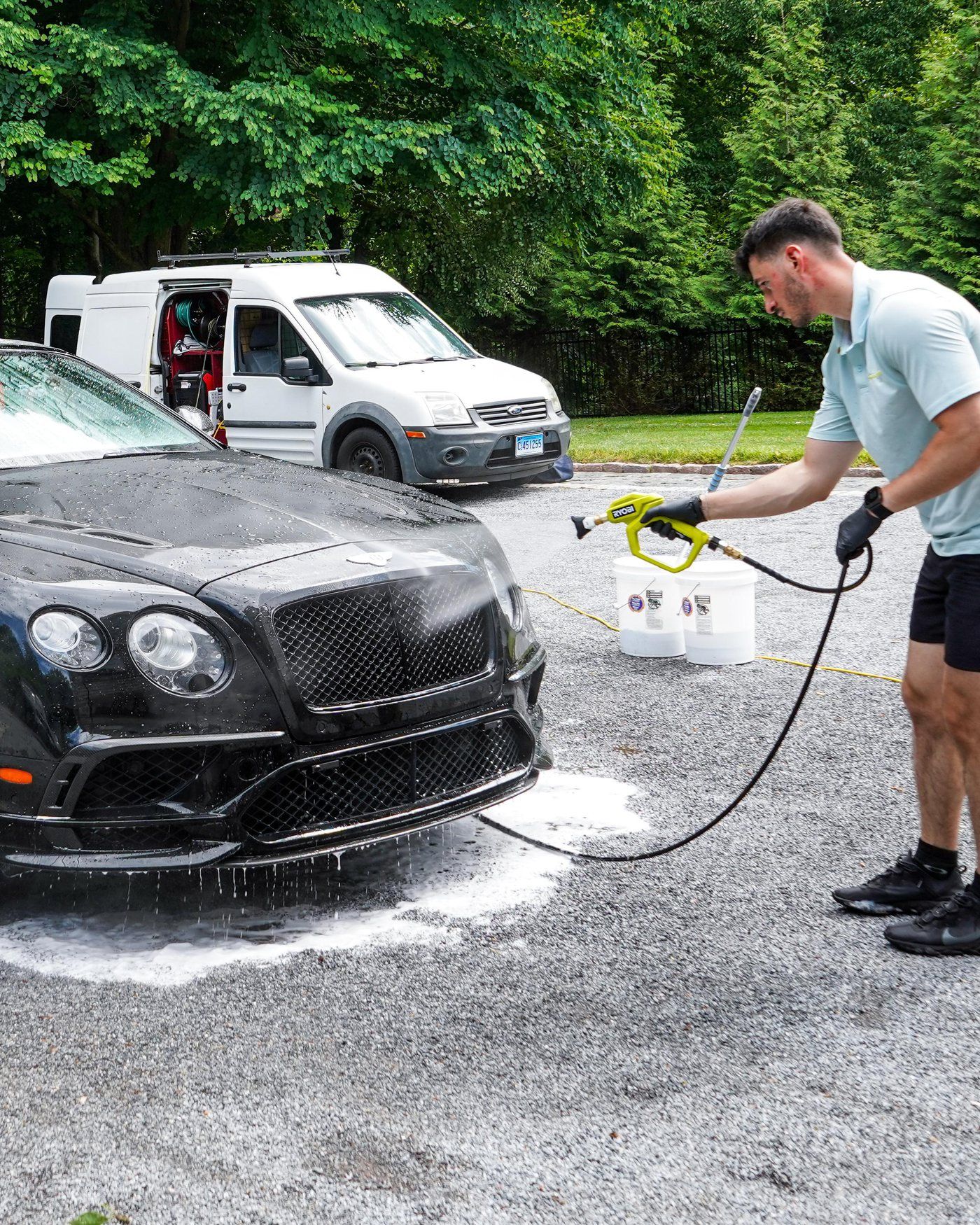 A detailer rinsing a black Bentley on a gravel drive, his white work van open behind him with the hose reel and buckets in view.