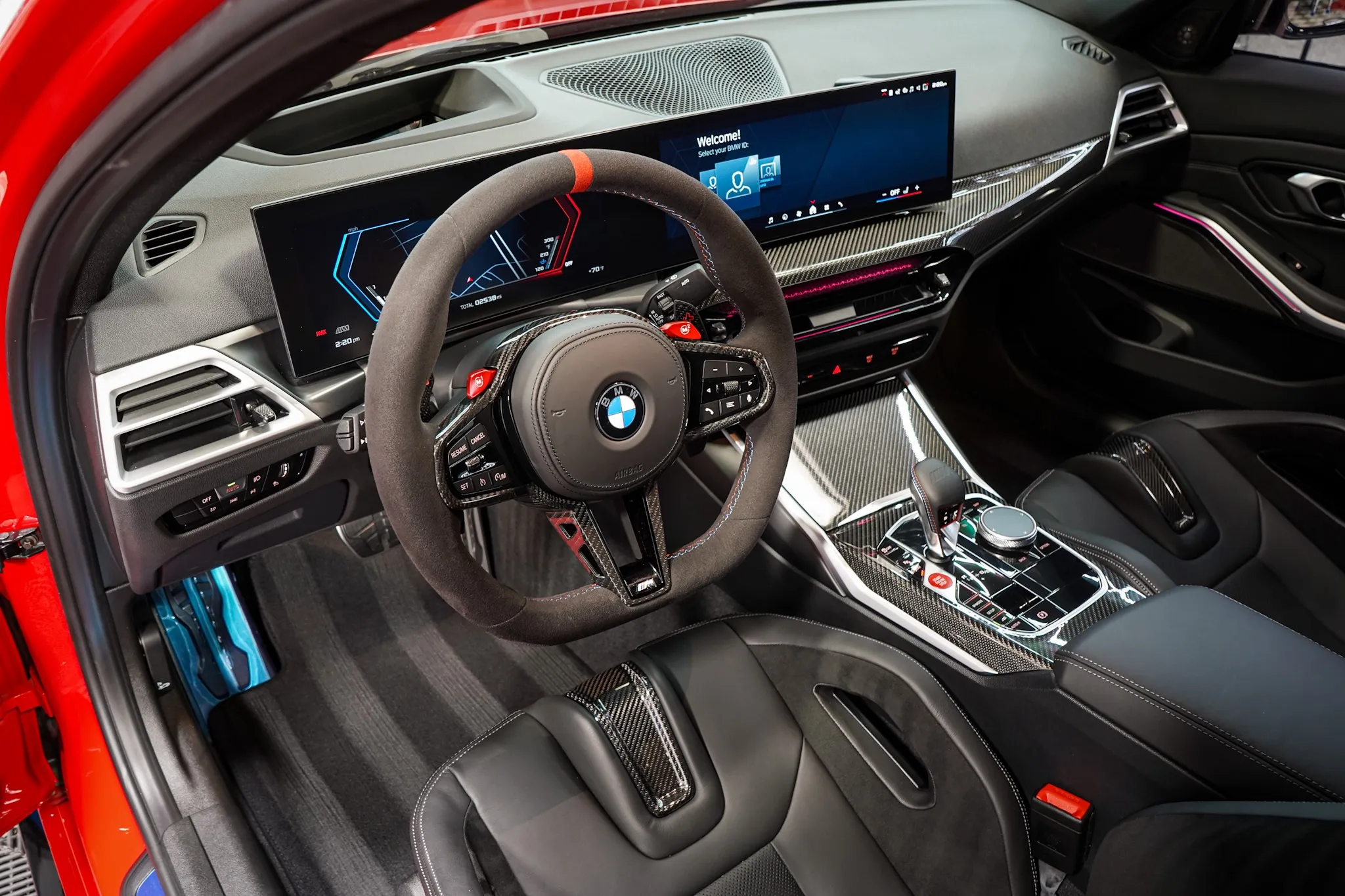 The driver's side of a BMW M interior: Alcantara steering wheel, carbon fiber trim and the two dashboard screens, seen through the open door.