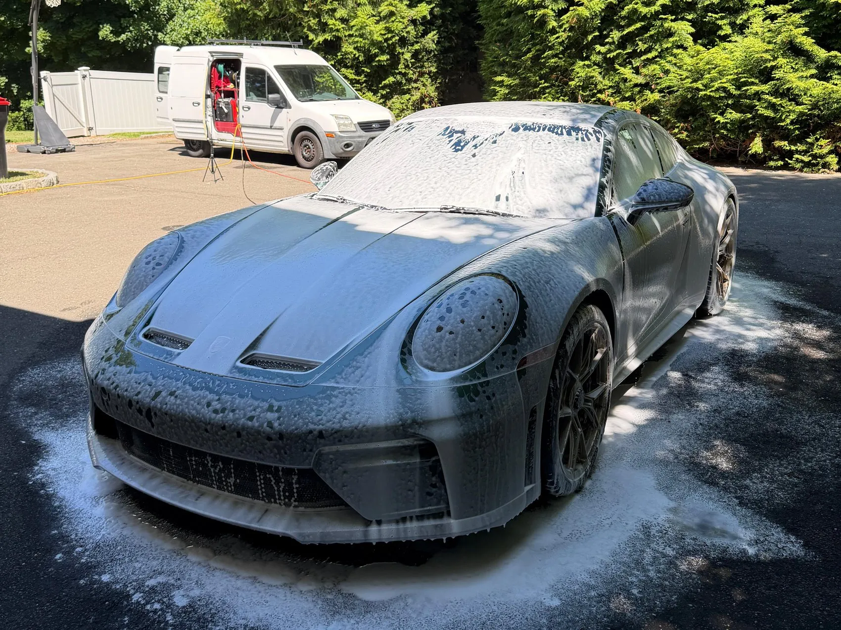 A Porsche 911 under snow foam on a driveway, with the white work van open behind it and the hose running across the tarmac.