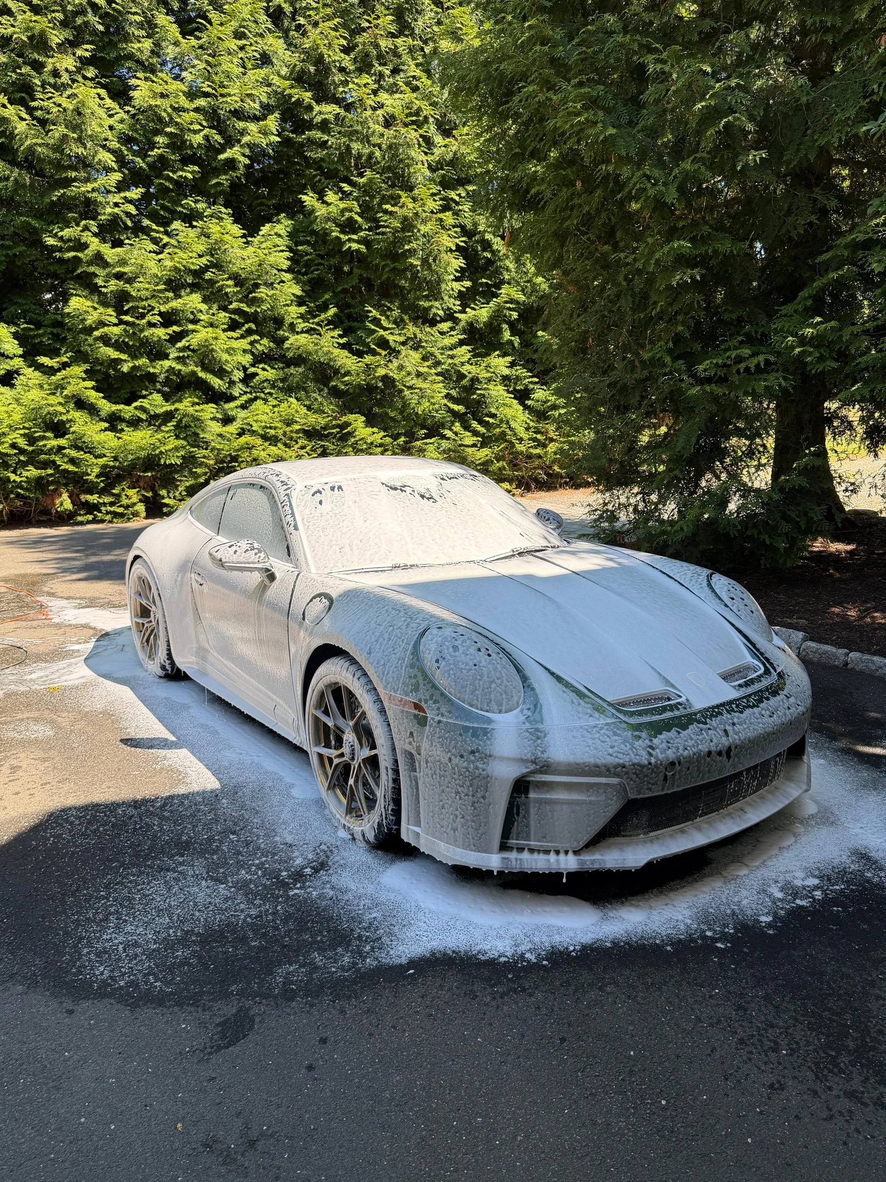 A Porsche 911 covered in white snow foam on a driveway, mid-wash.